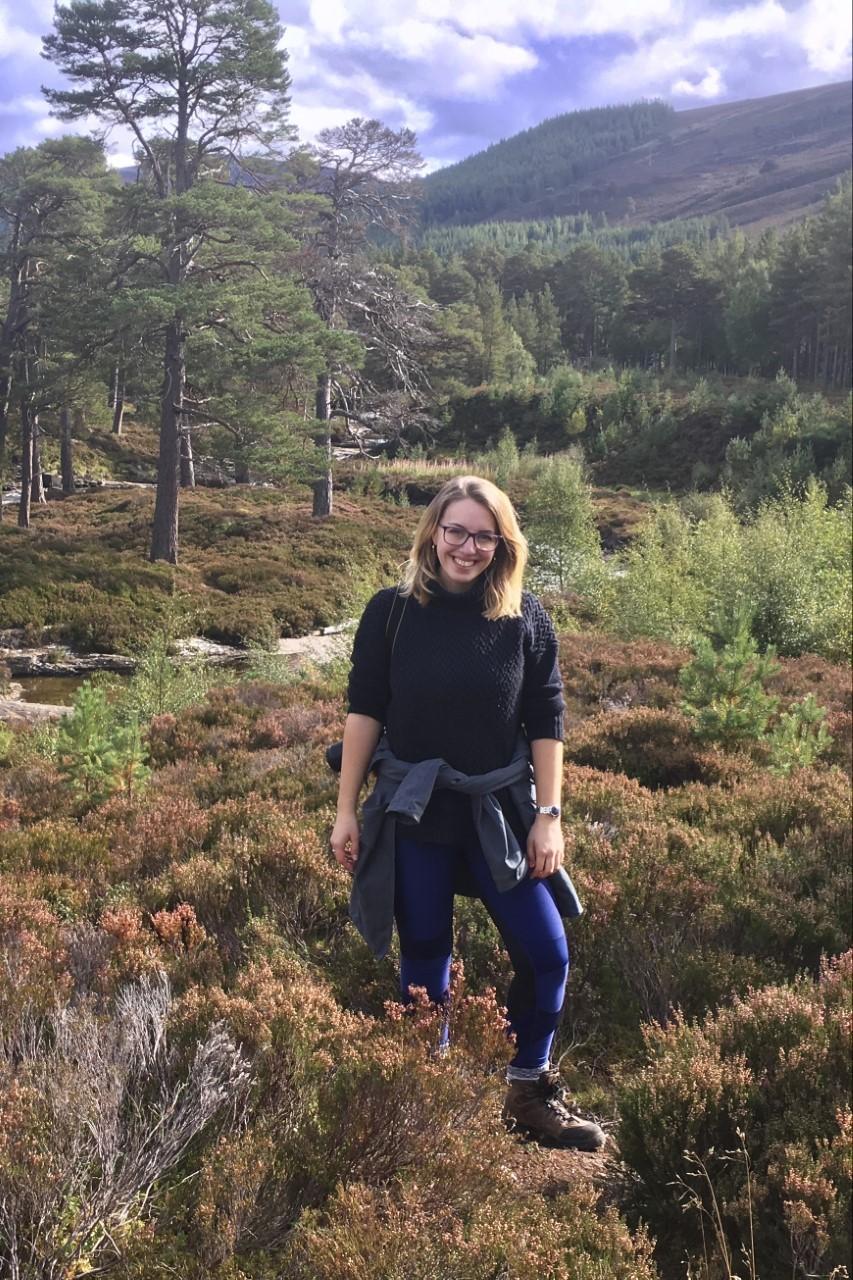 A picture of Alexandra Hoadley out walking in a glen. She is in mixed heather and pine wood, with a river behind and a mountain beyond that. She is smiling.